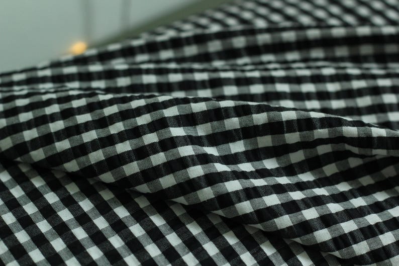 Op de afbeelding: Een stuk stof met een zwart-wit gingham ruitpatroon. Het materiaal heeft een uitgesproken gekreukelde, bobbelige textuur over het gehele oppervlak, wat zorgt voor zichtbare plooien en diepte in het geruite textielmateriaal.