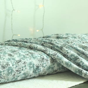 May include: A folded length of lightweight fabric featuring a soft sage green and white mottled background pattern overlaid with a repeating black line drawing of small, stylized flowers with thin petals and stems, creating a delicate visual texture.