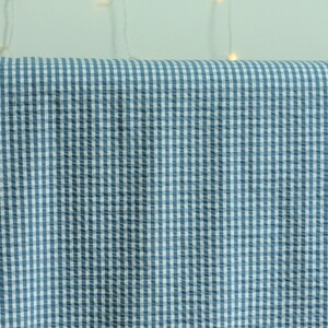 Op de afbeelding: Een stuk stof met een blauw-wit vichy-ruitpatroon. Het materiaal heeft een uitgesproken seersucker-textuur met een geplooid oppervlak, waardoor een raster van kleine vierkantjes over het gehele zichtbare deel van het textiel ontstaat.