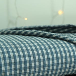 Op de afbeelding: Een gevouwen textiel met een blauw en wit vichy-ruitpatroon. De stof heeft een zichtbaar getextureerd oppervlak met een herhalend klein vierkant rasterontwerp. Het materiaal is gelaagd en vertoont een zachte plooiing en een consistent kleurenpalet.
