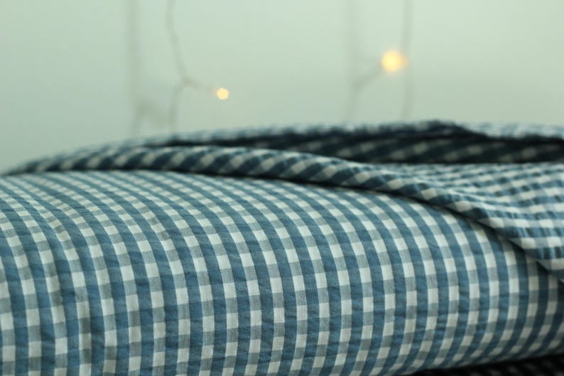 Op de afbeelding: Een gevouwen textiel met een blauw en wit vichy-ruitpatroon. De stof heeft een zichtbaar getextureerd oppervlak met een herhalend klein vierkant rasterontwerp. Het materiaal is gelaagd en vertoont een zachte plooiing en een consistent kleurenpalet.