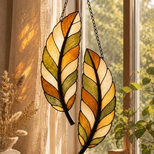 May include: Two hanging stained glass feather ornaments featuring a segmented design with alternating panels of amber, olive green, and cream-colored glass. Each feather is outlined in dark metal framing and suspended by a thin black metal chain.