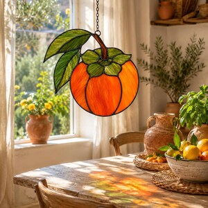 May include: A stained glass suncatcher shaped like a persimmon fruit with a vibrant orange body and green leaves, suspended by a metal chain. The glass features distinct dark lead lines outlining the segments of the fruit and the foliage.