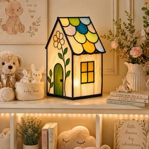 May include: A house-shaped stained glass lamp featuring a multi-colored scalloped roof, a green arched door, and a flower motif on the front panel. The lamp emits a warm glow through its translucent panels, which are framed in black metal.