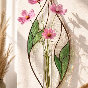 May include: A wall-mounted stained glass leaf-shaped vase featuring a clear glass test tube holder. The frame is made of dark metal with textured clear and green glass panels. Three pink cosmos flowers with yellow centers are placed inside.