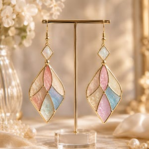 May include: A pair of gold-toned dangle earrings featuring a diamond-shaped frame filled with segmented, iridescent panels in shades of pastel pink, blue, and cream. Each earring includes a small diamond-shaped stud connected to the larger drop.