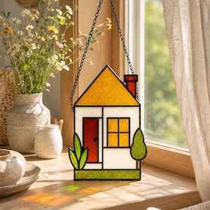 May include: A stained glass suncatcher shaped like a house with a yellow roof, white walls, a red door, and a yellow window. The house features a red chimney, a green base, and green foliage on both sides. It hangs from a black metal chain.