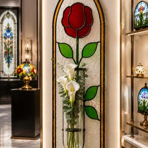 May include: A tall, arched stained glass wall hanging featuring a red rose and green leaves on a textured white background. A clear glass cylindrical vase is attached to the centre, holding three white calla lilies and green eucalyptus stems.