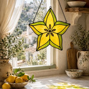 May include: A star-shaped stained glass suncatcher featuring a vibrant yellow centre with dark brown seed-like accents, surrounded by a bright green border. The piece is suspended by a thin black metal chain against a window pane.