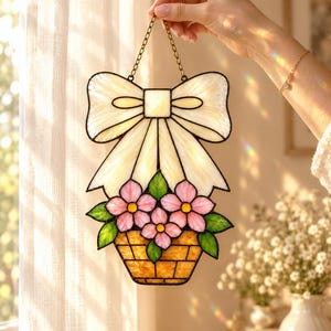 May include: A stained glass suncatcher featuring a cream-coloured bow above a basket filled with pink flowers and green leaves. The piece is constructed with dark metal soldering and hangs from a thin gold-toned metal chain.