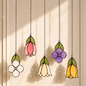 May include: Five stained glass flower ornaments hanging from thin metal chains. The flowers feature white, pink, cream, purple, and yellow petals with green leaves, all outlined in dark metal solder, creating a colourful floral display.