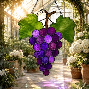 Puede incluir: Un atrapasoles de vidrieras con forma de racimo de uvas moradas con dos grandes hojas verdes y un tallo marrón, suspendido por una cadena de metal negro. El vidrio presenta una superficie texturizada que atrapa la luz, creando un brillo morado.