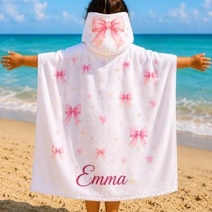 Pode incluir: Um poncho de toalha branco com capuz apresentando um padrão repetitivo de laços cor-de-rosa, pequenos corações cor-de-rosa e brilhos dourados. Um grande laço cor-de-rosa está estampado no capuz. O nome Emma está escrito em letras cursivas.
