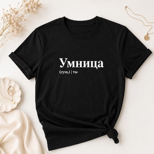 Op de afbeelding: Een zwart t-shirt met korte mouwen en witte tekst op de borst. De tekst luidt "Умница" met daaronder "(сущ.) | ты". Het shirt heeft een ronde hals en opgerolde mouwen, wat zorgt voor een strak en eenvoudig ontwerp.