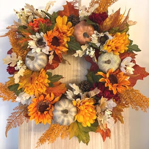 May include: A decorative wreath featuring artificial sunflowers, orange and red autumn leaves, pinecones, white berries, and three small pumpkins. The pumpkins are textured and light-colored, nestled among vibrant orange, yellow, and deep red floral elements.