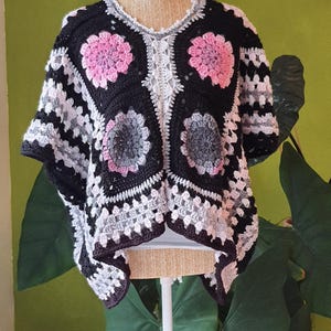 Peut inclure: Un poncho crocheté à la main en noir, gris et blanc, orné de motifs floraux roses et gris. Le poncho est présenté sur un mannequin sur fond vert, mettant en valeur son design ouvert et ses points texturés.