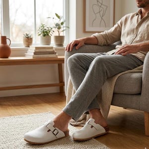 Könnte beinhalten: Weiße Lederclogs mit Schnallenverschluss und Korkfußbett. Die Clogs werden in Innenräumen auf einem hellen Teppich gezeigt. Das Bild zeigt eine Person, die in einem grauen Sessel sitzt.