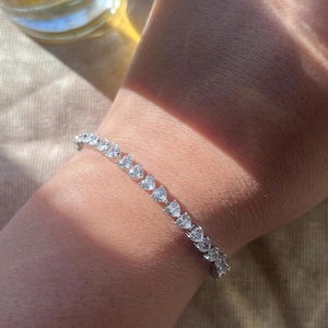 Peut inclure: Un bracelet en argent orné de pierres précieuses en forme de cœur. Le bracelet est porté sur un poignet, les pierres précieuses captant la lumière. L'arrière-plan est de couleur neutre, mettant en valeur le bijou.