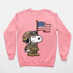 May include: Pink sweatshirt featuring a cartoon Snoopy character dressed as a soldier, wearing a helmet and holding an American flag. The design includes military gear and patriotic details, perfect for a themed gift or casual wear.