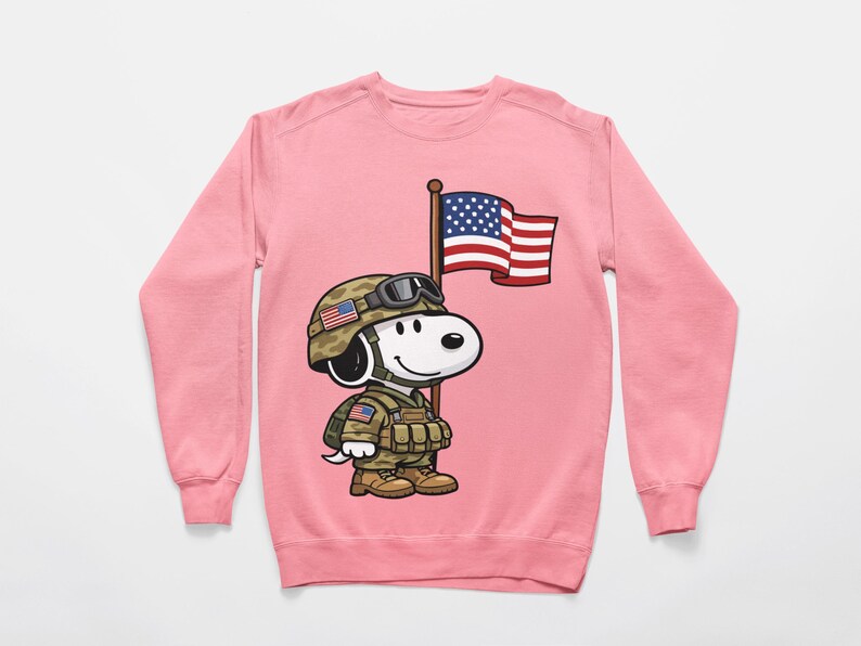 May include: Pink sweatshirt featuring a cartoon Snoopy character dressed as a soldier, wearing a helmet and holding an American flag. The design includes military gear and patriotic details, perfect for a themed gift or casual wear.