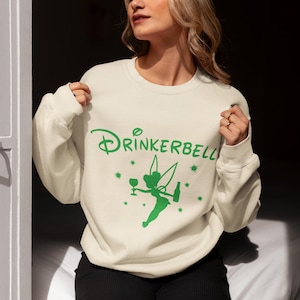 May include: Cream-colored sweatshirt with the word "Drinkerbell" in green script, featuring a green graphic of a fairy holding a wine glass and bottle. The design includes stars. The sweatshirt is a casual, comfortable garment.
