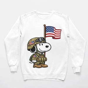 May include: White sweatshirt featuring a cartoon Snoopy character dressed as a soldier, wearing a camouflage helmet and uniform. Snoopy is holding a flagpole with an American flag. The design is a patriotic theme.