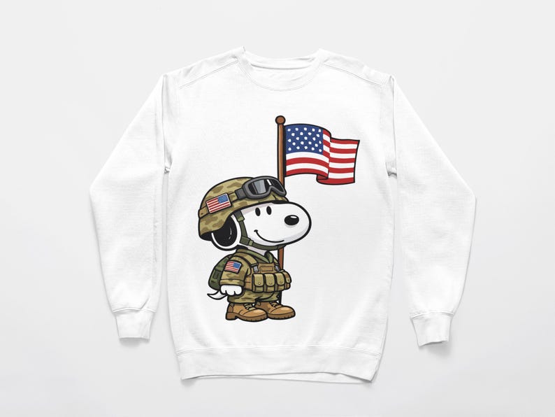 May include: White sweatshirt featuring a cartoon Snoopy character dressed as a soldier, wearing a camouflage helmet and uniform. Snoopy is holding a flagpole with an American flag. The design is a patriotic theme.