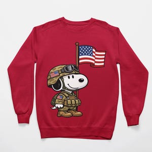 May include: A red sweatshirt featuring a cartoon character in military attire, complete with a helmet, uniform, and an American flag. The character is holding a flagpole with the American flag. The design is centered on the front of the sweatshirt.