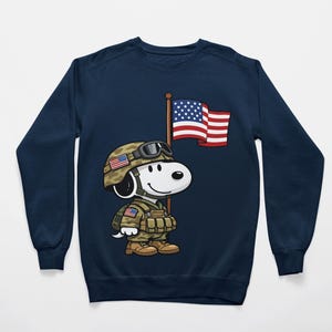 May include: Navy blue sweatshirt featuring a cartoon Snoopy character dressed as a soldier, wearing a helmet with an American flag design. Snoopy is holding a flagpole with the American flag. The design is centered on the front of the sweatshirt.