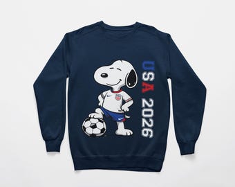 Może przedstawiać: Granatowa bluza z nadrukiem Snoopy'ego w stroju piłkarskim USA, pozującego z piłką nożną. Tekst "USA 2026" jest wydrukowany pionowo. Projekt wykorzystuje kolory czerwony, biały i niebieski.