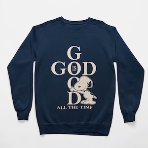 May include: Navy blue sweatshirt featuring the phrase "GOD IS GOOD ALL THE TIME" in a cream color. The design includes a Snoopy character. The long-sleeved top is a casual wear item.