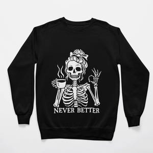 May include: Black sweatshirt with a white graphic of a skeleton woman with a bun, holding a coffee cup and making an "okay" hand gesture. The text "NEVER BETTER" is below the graphic. A casual, humorous design.
