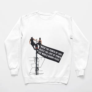 May include: A white long-sleeve sweatshirt featuring a black graphic print of two figures perched on a pole. The graphic includes a black rectangular sign with the white text: WHEN THE POWER OF LOVE BEATS THE LOVE OF POWER THE WORLD KNOWS PEACE.