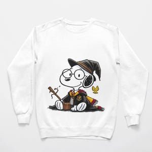May include: White sweatshirt featuring a cartoon Snoopy character dressed as a wizard, wearing a black hat, glasses, and a Hogwarts scarf. Snoopy is holding a wand and sitting on a white surface. A golden emblem and a small yellow bird are also visible.