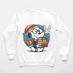 May include: White sweatshirt featuring a cartoon Snoopy character wearing glasses, a scarf, and a hat, carrying books labeled "MAGIC." An owl flies nearby against a blue background with stars.