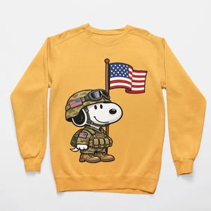 May include: Mustard yellow sweatshirt featuring a cartoon Snoopy character dressed as a soldier, complete with a helmet, military uniform, and holding a US flag. The design is a patriotic tribute, perfect for casual wear.