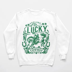 Puede incluir: Sudadera blanca con un gráfico verde. El gráfico presenta un vaquero montando un caballo, las palabras "Feeling Lucky Cowboy", tréboles y herraduras. El diseño está centrado en la parte delantera de la parte superior de manga larga.