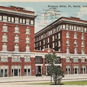 May include: Vintage postcard featuring the Goldman Hotel in Ft. Smith, Ark. The postcard shows a detailed illustration of the brick hotel with multiple windows and a white trim. The sky is light blue, and trees are visible on either side of the building.