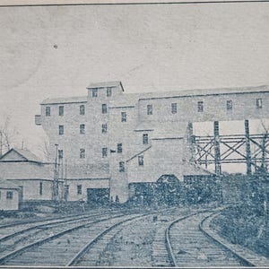 May include: Vintage postcard featuring the Russellville Anthracite Coal Mining Co. in Russellville, Arkansas. The black and white image shows the coal mine buildings, train tracks, and a smokestack. The postcard has text that reads "Daily capacity, 500 Tons."
