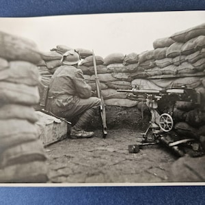 May include: Black and white photograph of a soldier in a trench, seated with a rifle. A machine gun is positioned nearby, with sandbags providing cover. The scene depicts a moment of rest or observation during wartime.