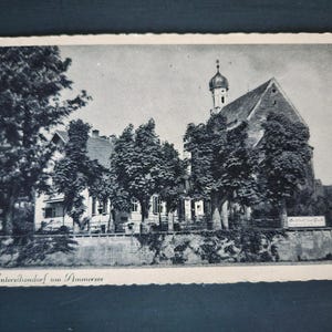 May include: Black and white vintage postcard featuring a church with a tall steeple and a pitched roof. Trees line the front of the building. The postcard has a white border and the text "Unterschondorf am Ammersee" at the bottom.