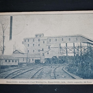 May include: Vintage black and white postcard depicting the Russellville Anthracite Coal Mining Co. in Russellville, Arkansas. The image shows the coal mining facility with multiple buildings, a tall smokestack, and railroad tracks. The text at the bottom reads "Russellville Anthracite Coal Mining Co., Russellville, Ark, Daily capacity, 800 Tons."