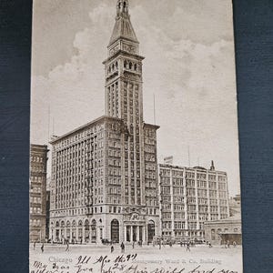 May include: Vintage black and white postcard of the Montgomery Ward & Co. Building in Chicago, Illinois. The tall building has a prominent clock tower and is surrounded by other buildings and people. Text on the postcard reads "Chicago Ill After the 9th".