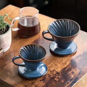 Op de afbeelding: Twee blauwe keramische koffiefilters met interne verticale ribbels en een handvat, naast een heldere glazen karaf gevuld met donkerbruine vloeistof. De filters hebben een diepblauw glazuur met donkerbruine randen aan de bovenkant.