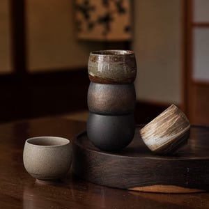 Può includere: Una collezione di tazze da tè in ceramica artigianali in vari colori naturali. Le tazze sono impilate e disposte su una superficie di legno scuro. Le tazze da tè hanno un aspetto rustico e strutturato, suggerendo un'attenzione ai materiali naturali e al design minimalista.
