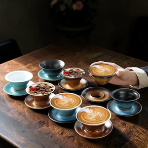 Puede incluir: Una colección de tazas y platillos de cerámica en varios colores, incluyendo azul, marrón y blanco, dispuestos sobre una mesa de madera. Algunas tazas contienen café con arte latte, otras postres.