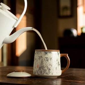 Pode incluir: Uma caneca de cerâmica branca com uma alça e borda marrom, com um desenho de floresta de bambu. Uma chaleira branca despeja líquido na caneca. Uma tampa de madeira está ao lado da caneca em uma mesa de madeira. A caneca tem aproximadamente 10 cm de altura.