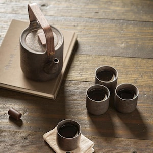 Könnte beinhalten: Ein rustikales Teeservice mit einer dunkelgrauen Teekanne mit Holzgriff, die auf einem Buch liegt. Drei passende Tassen und ein kleines Holzstück sind auf einer Holzoberfläche angeordnet, was eine ruhige, minimalistische Ästhetik schafft.
