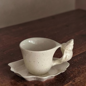 Peut inclure: Un ensemble tasse et soucoupe en céramique blanc cassé moucheté. La tasse présente une anse en forme de coquillage et repose sur une soucoupe assortie à bord festonné. L'ensemble a une esthétique rustique et artisanale.