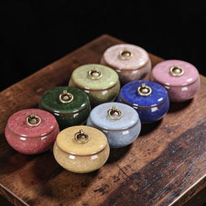 Może przedstawiać: Kolekcja ośmiu małych, ceramicznych słoiczków z pokrywkami. Słoiczki są w różnych kolorach, w tym czerwonym, żółtym, zielonym, niebieskim i różowym. Każdy słoiczek ma złoty uchwyt w kształcie pierścienia na pokrywce. Słoiczki są ułożone na drewnianej powierzchni.